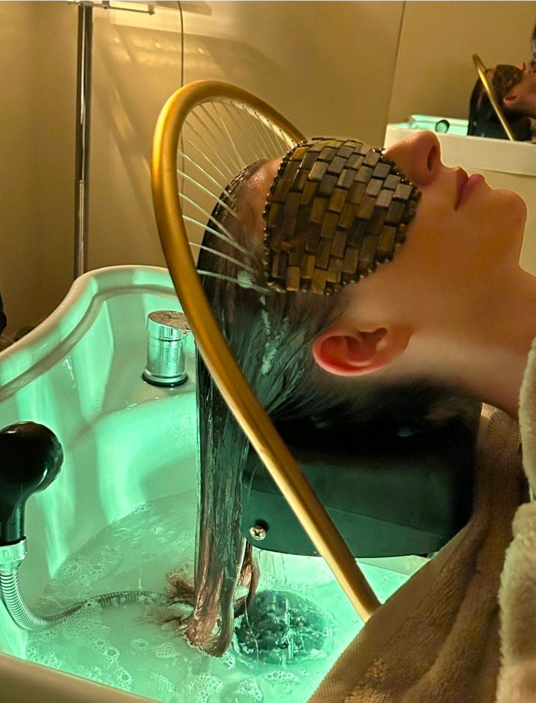 Cliente allongée, la nuque au-dessus du bac de head spa, sous les fils d'eau de l'arceau doré, un masque en bois sur les yeux