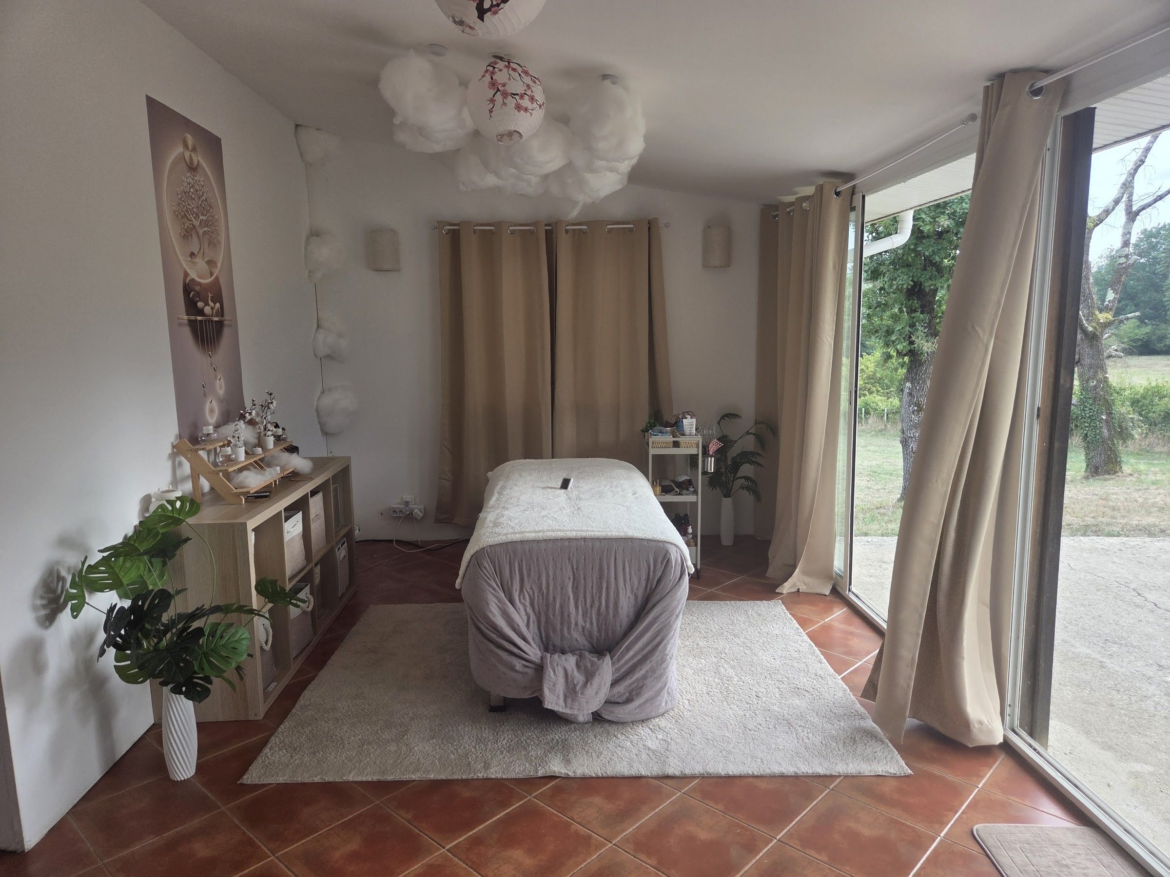 La salle de soin en journée : table de soin, plafond de nuages et lanternes, baie vitrée sur le jardin
